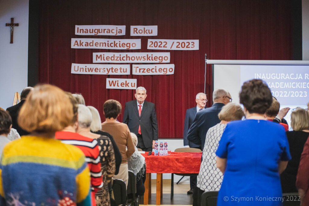 Na zdjęciu przedstawioni są studenci Miedzichowskiego UTW wraz z zaproszonymi gośćmi. Na drugim planie przedstawieni są dwie osoby, którzy rozpoczynają rok akademicki 2022/2023. Na trzecim planie jest tło czerwone z napisem Inauguracja Roku Akademickiego 2022/2023 Miedzichowskiego Uniwersytetu Trzciego Wieku.