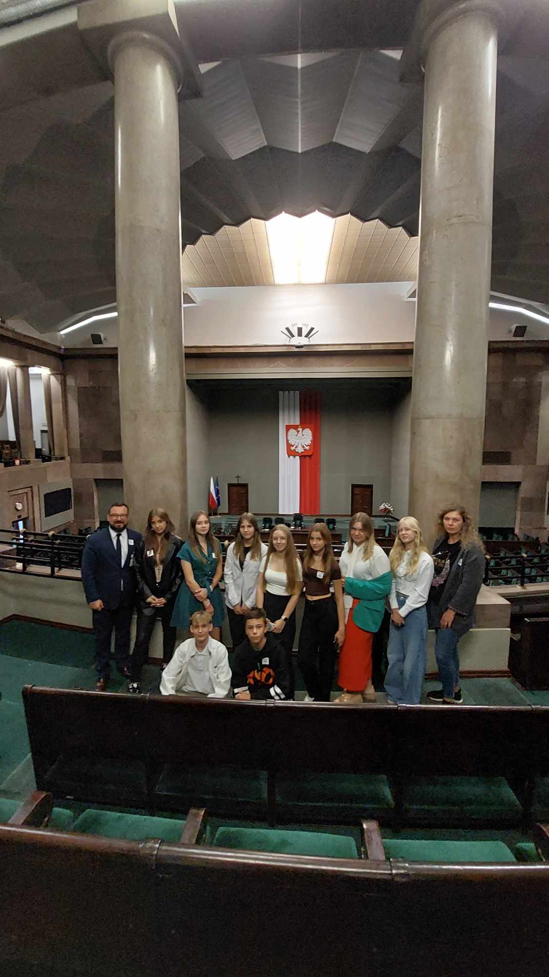 Zdjęcie ukazuję Młodzieżową Radę Gminy w&nbsp;tle sali konferencyjnej w&nbsp;Sejmiku. Za młodzieżową radą widaczna jest sala wraz z&nbsp;udekorowaniem. 