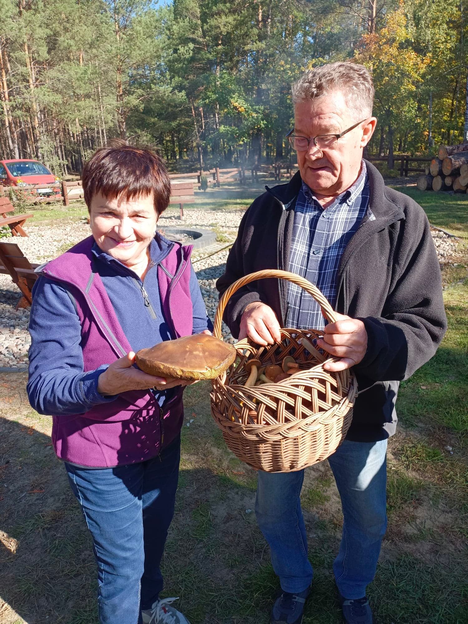 Na zdjęciu pozują dwie osoby trzymające koszyk z grzybami. Po lewej stronie kobieta w brązowych włosach oraz niebieskiej bluzie i fioletowej kamizelce. Po prawej stronie stoi mężczyzna w okularach i siwych włosach. Ubrany jest w niebieską koszulę w kratkę i czarną bluzę.