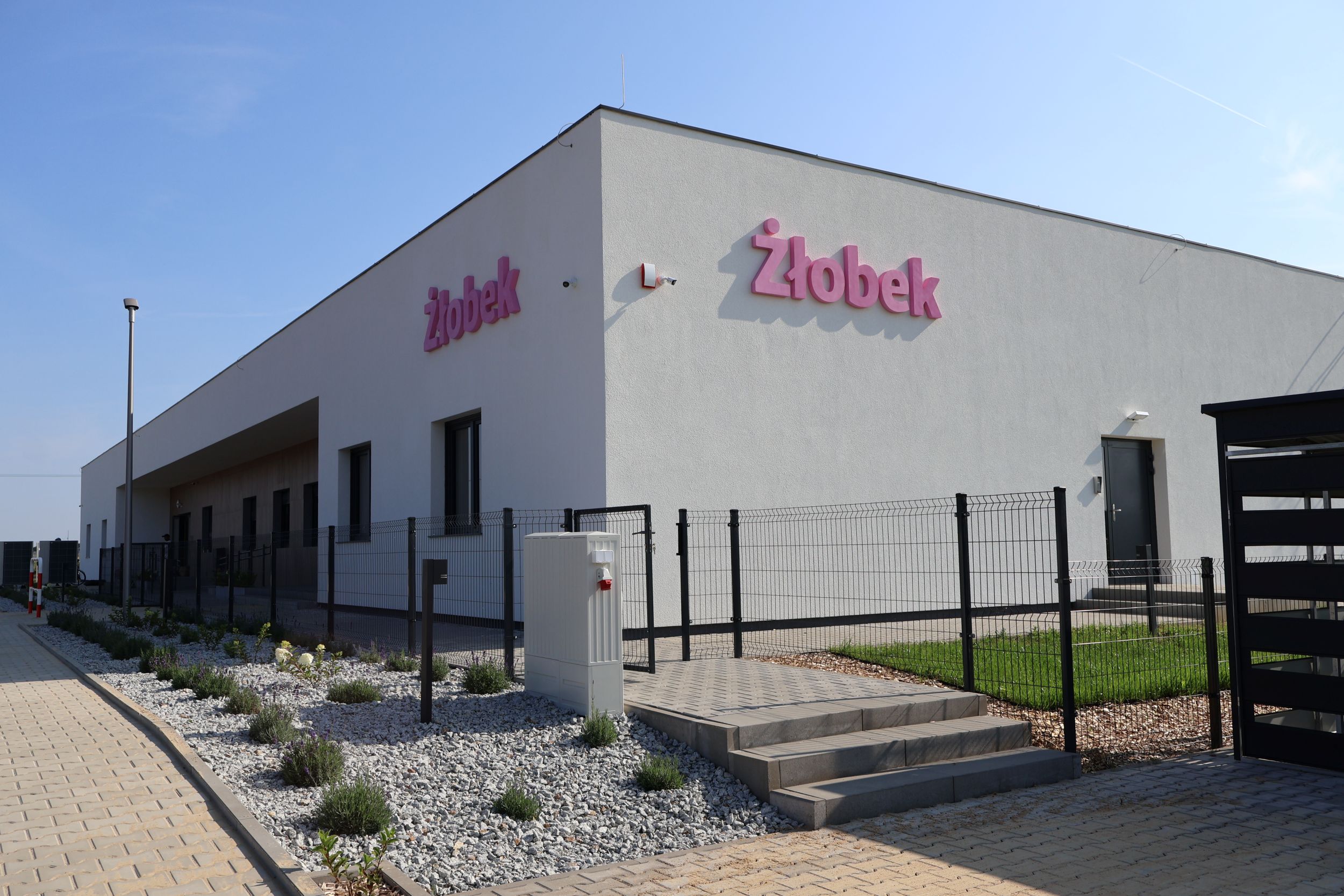 Grafika przedstawia budynek w&nbsp;kolorze jasnego szarego z&nbsp;napisem r&oacute;żowym złobek. Przed budynkiem znajduje się ogrodzenie oraz&nbsp;zielona powierzchnia.