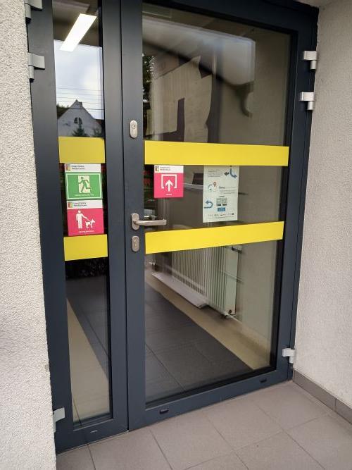 Wejście urzędu. Drzwi są przezroczystę i&nbsp;oklejoną ż&oacute;łtą taśmą.ę i&nbsp;oklejoną ż&oacute;łtą taśmą oraz&nbsp;oznaczone tabliczkami dla os&oacute;b z&nbsp;niepełnosprawnościami.