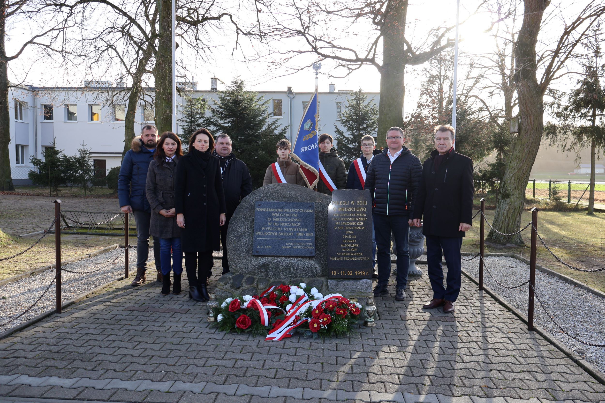 Wsp&oacute;lne zdjęcie przy pomniku im. Powstańc&oacute;w Wielkopolski w&nbsp;Miedzichowie od lewej strony Rada Sołecka wraz z&nbsp;Sołtysem, Sekretarz Gminy Miedzichowo, sztandar szkolny, Dyrektor Szkoły w&nbsp;Miedzichowie i&nbsp;W&oacute;jt Gminy Miedzichowo. Wszyscy są ubrani w&nbsp;ciemnych kolorach.
