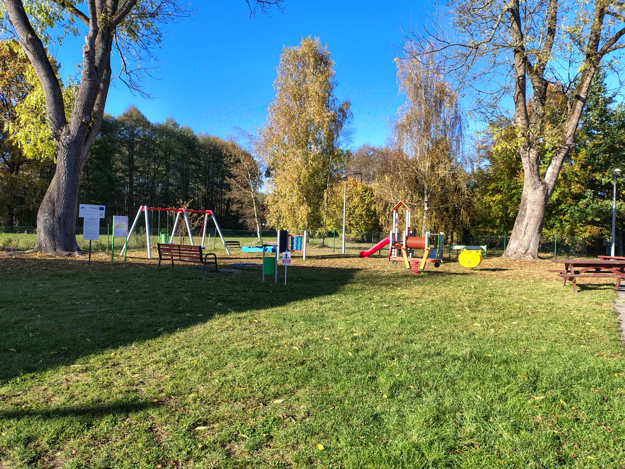 Zdjęcie przedstawia teren placu zabaw, między drzewami na&nbsp;zielonej trawie.