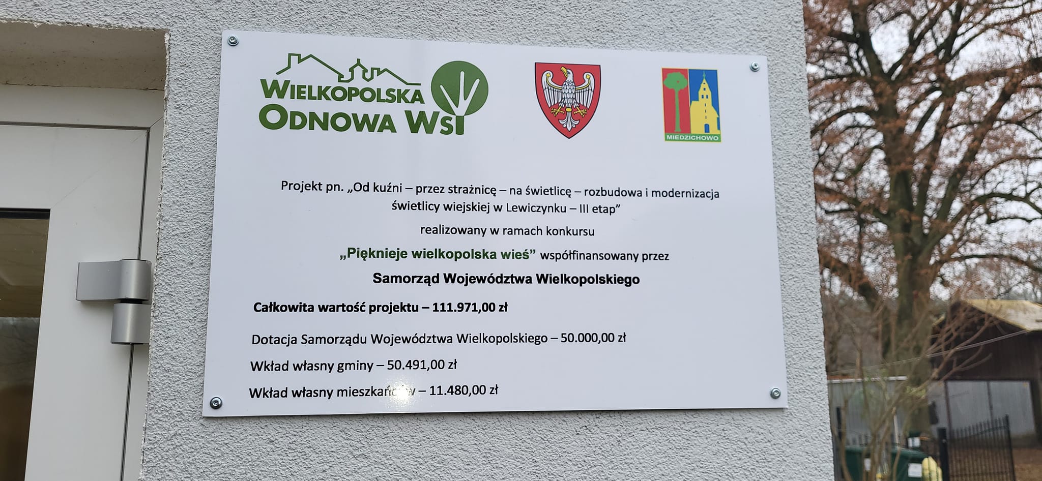 tablica "Projekt pn. "Od kuźni-przez strażnicę -na świetlicę - rozbudowa i modernizacja świetlicy wiejskiej w Lewiczynku - III etap" realizowany w ramach konkursu "Pięknieje wielkopolska wieś" współfinansowany przez Samorząd Województwa Wielkopolskiego Całkowita wartość projektu - 111.971,00 złDotacja Samorządu Województwa Wielkopolskiego - 50.000,00 zł Wkład własny gminy -50.491,00 zł wkład własny mieszkańców 11.480,00 zł.