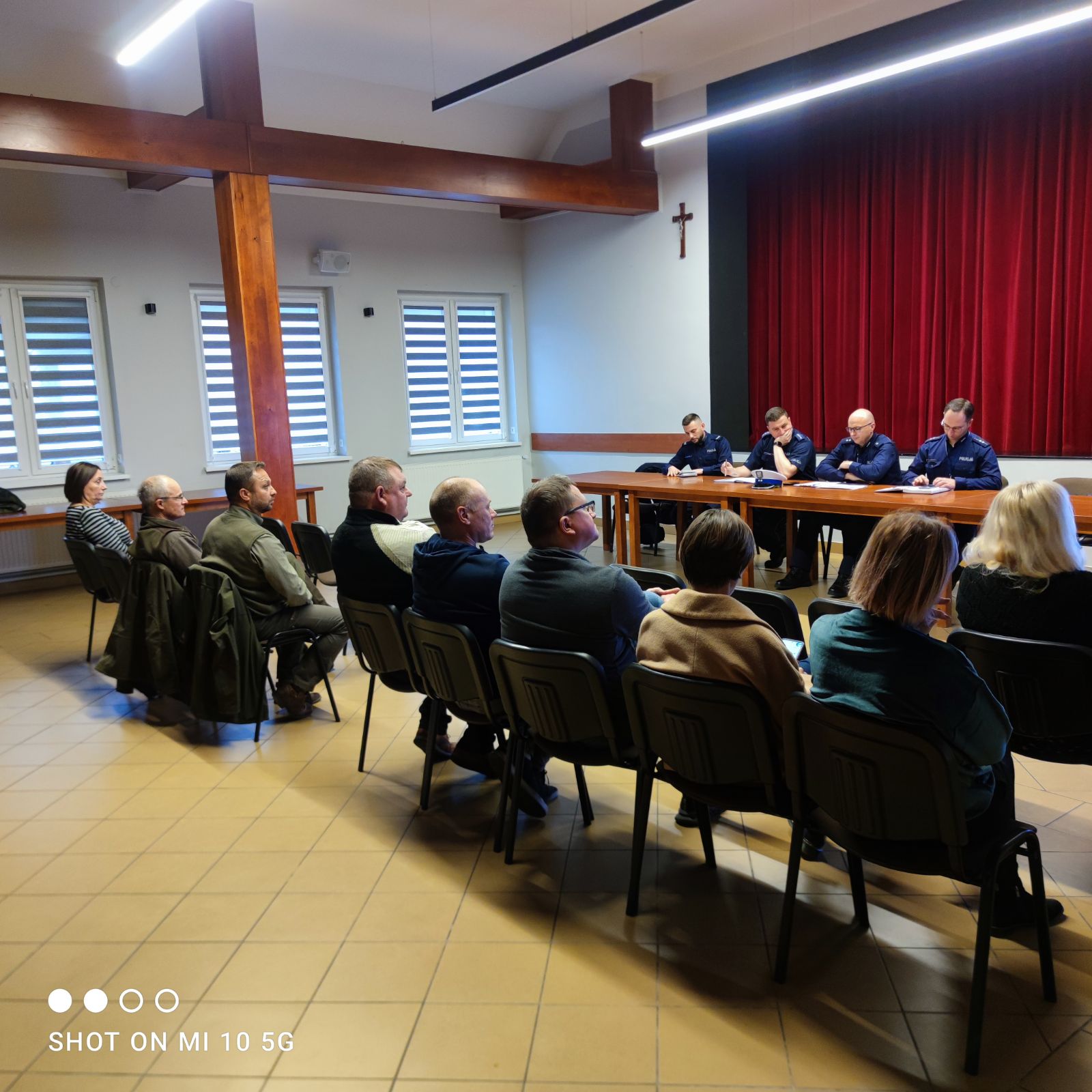 Pierwszy plan przedstawia uczestnik&oacute;w spotkanie, kt&oacute;rzy siedzą na&nbsp;ciemnych krzesłach prostopadle do okna na&nbsp;przeciwko funkcjonariuszy. Policja umiejscowiona jest przed uczestnikami siedzą przy stole w&nbsp;ciemnym kolorze.